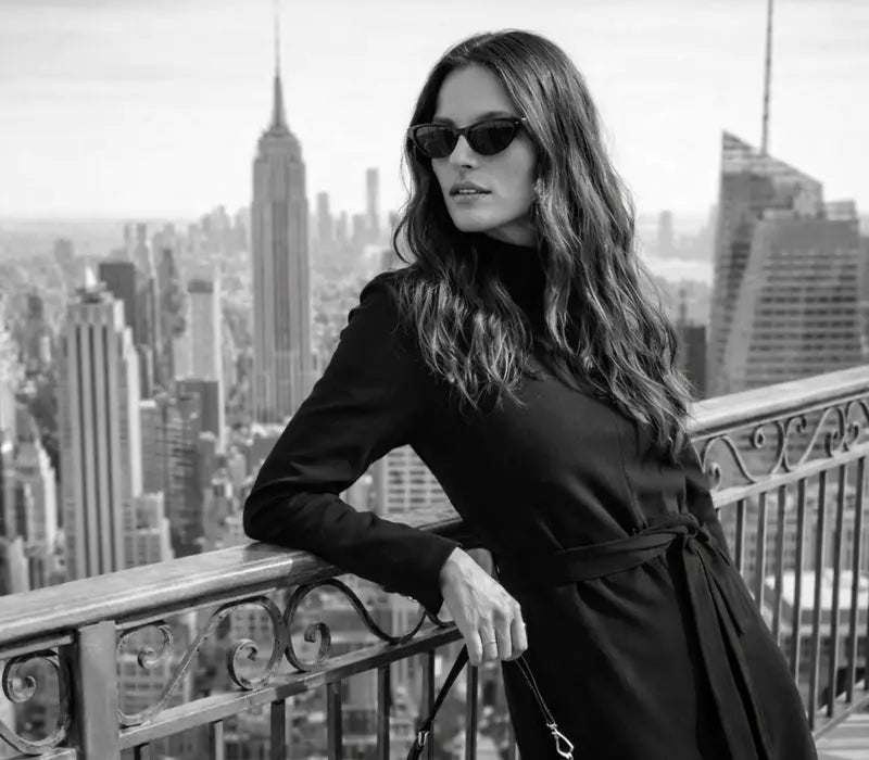 Woman wearing black long-sleeve jersey dress standing on balcony with New York City skyline background