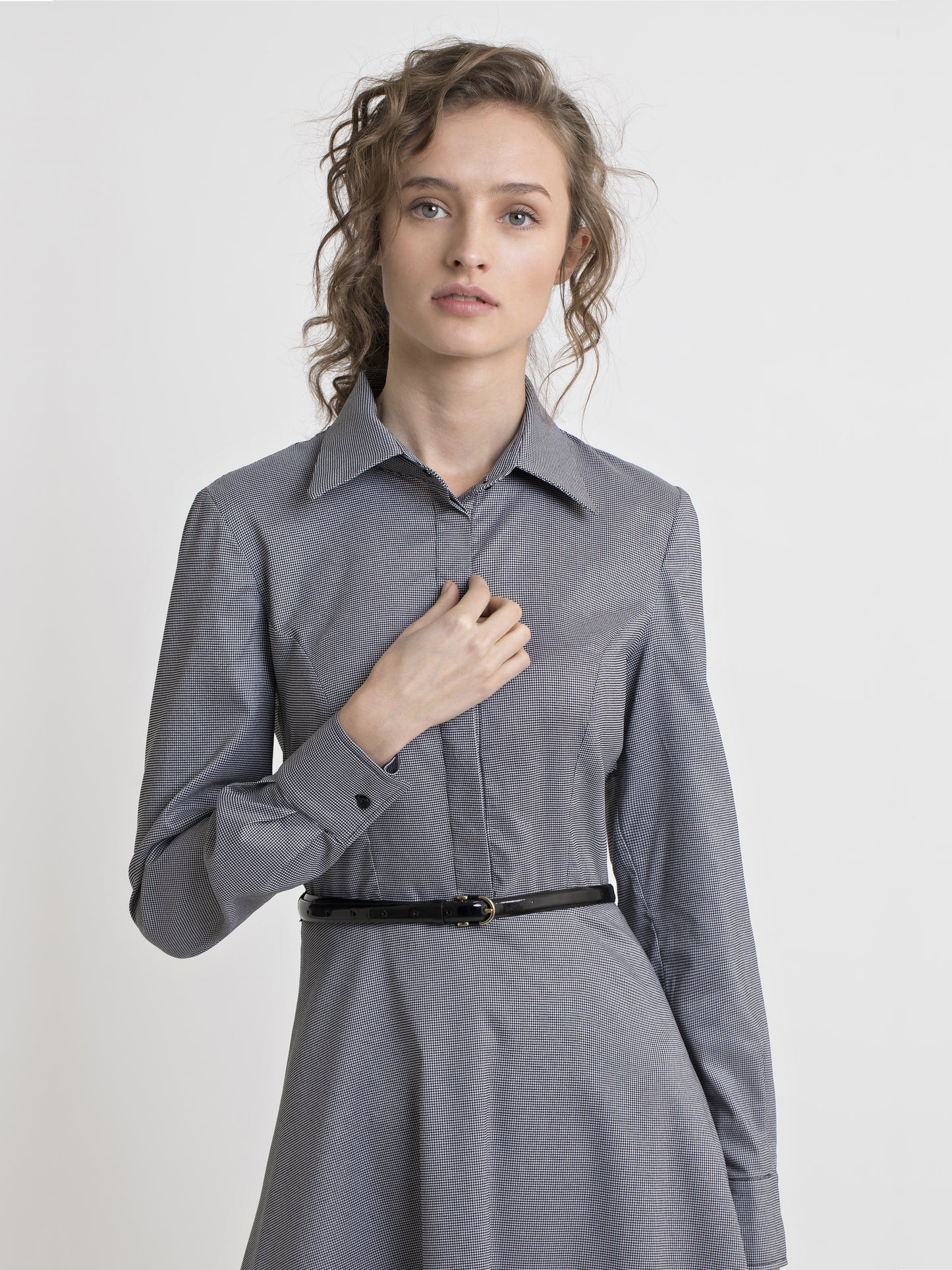 Upper body view of a female model, wearing a semi fitted black and white check shirt dress, with covered placket and a patent leather black narrow belt. From the RÉZO women's collection.