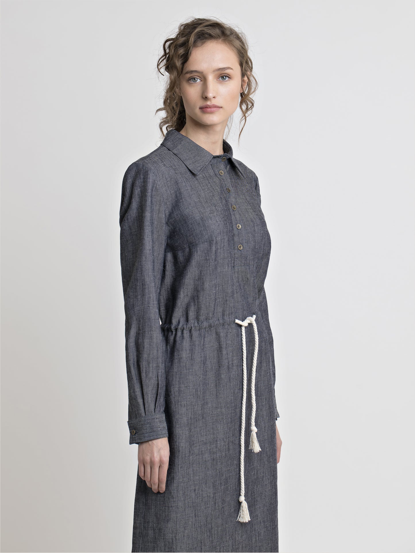 3/4 view of a female model wearing long sleeve straight cut denim shirt dress with drawstring inserted belt. From the RÉZO women's collection.