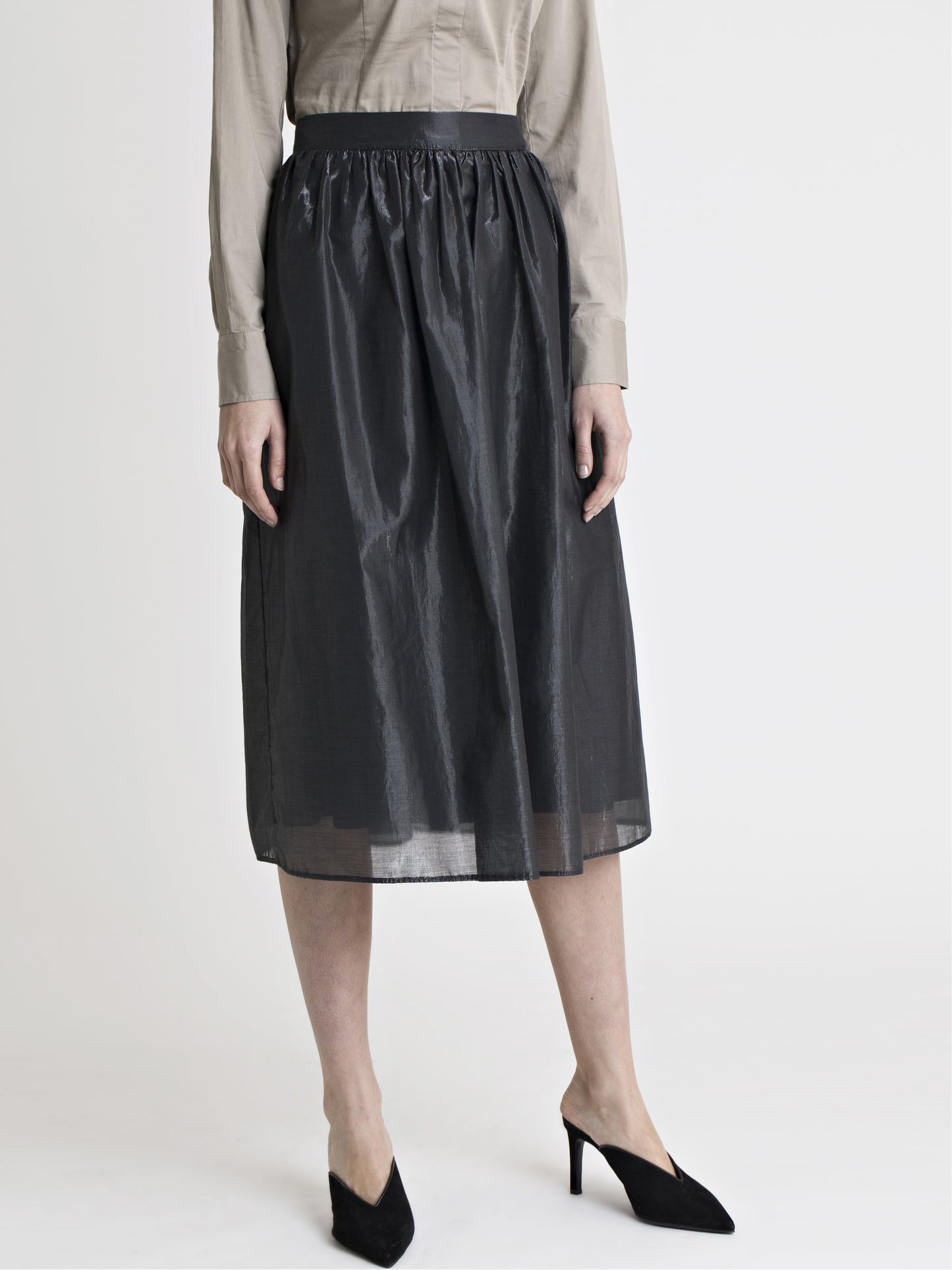 Lower body front view of a female model wearing a sand color dress shirt, black high heel slip on shoes, and a below the knee glossy black gathered skirt, from the RÉZO women's collection.