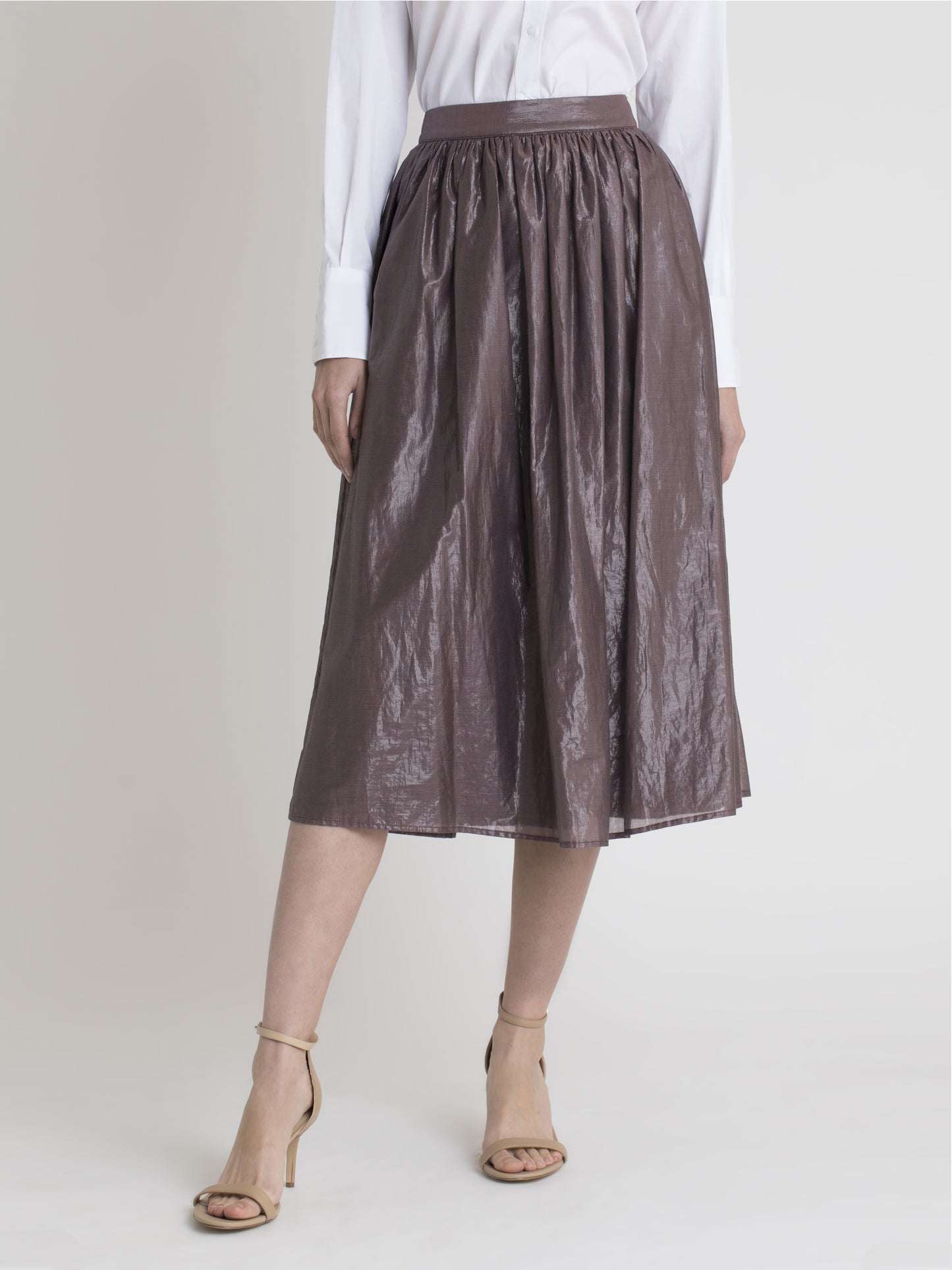 Lower body front view of a female model wearing a white dress shirt, nude high heel sandals, and a below the knee glossy earth color gathered skirt from the RÉZO women's collection.