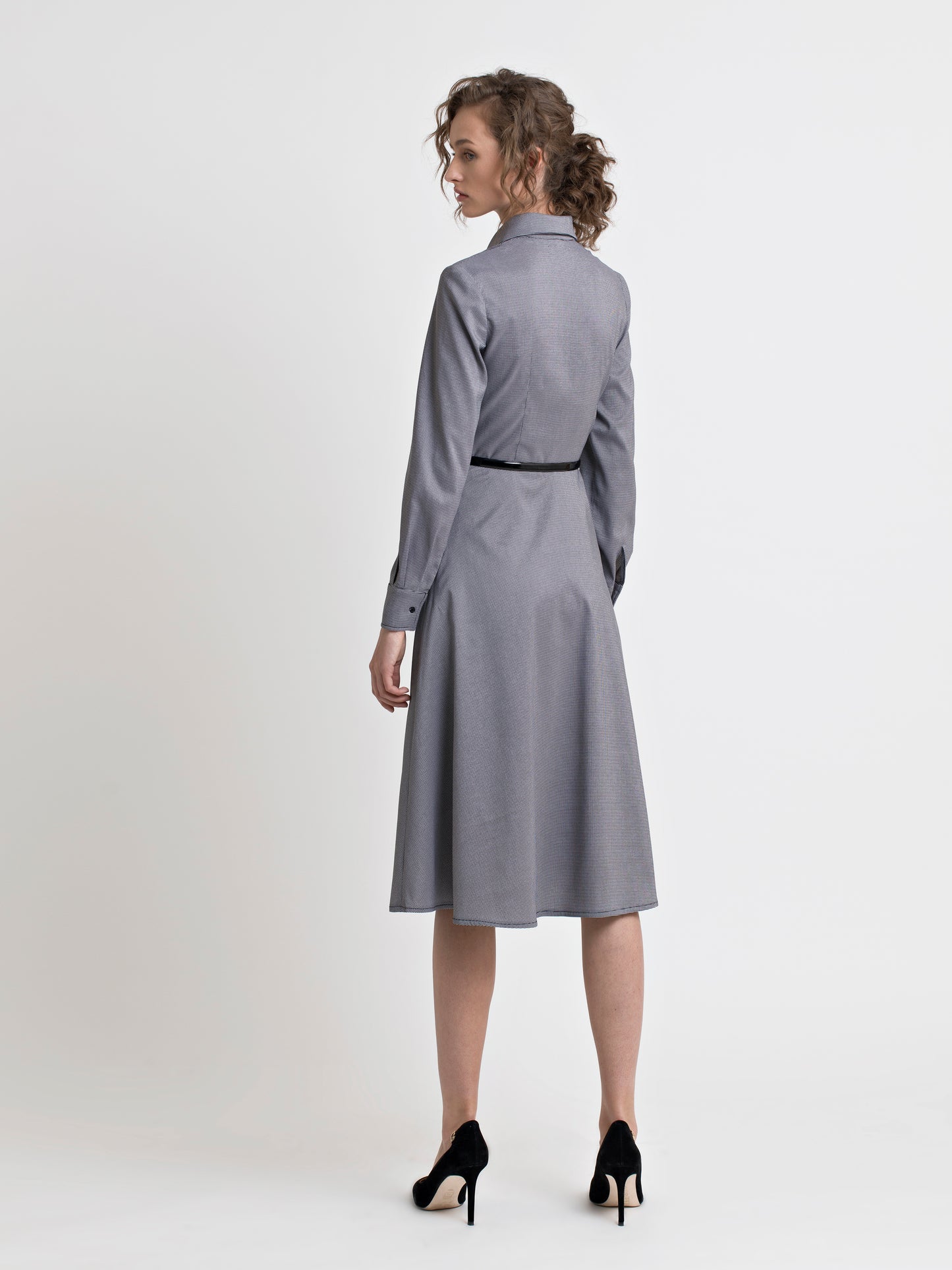 Back view of a female model, wearing high heel black shoes, and a semi fitted black and white check shirt dress with a patent leather black narrow belt. From the RÉZO women's collection.